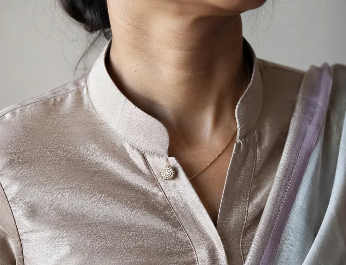 Close up of an Indian woman wearing a mandarin collar raw silk saree blouse with a statement button and delicate gold chain, highlighting a modern blouse design for 2026.