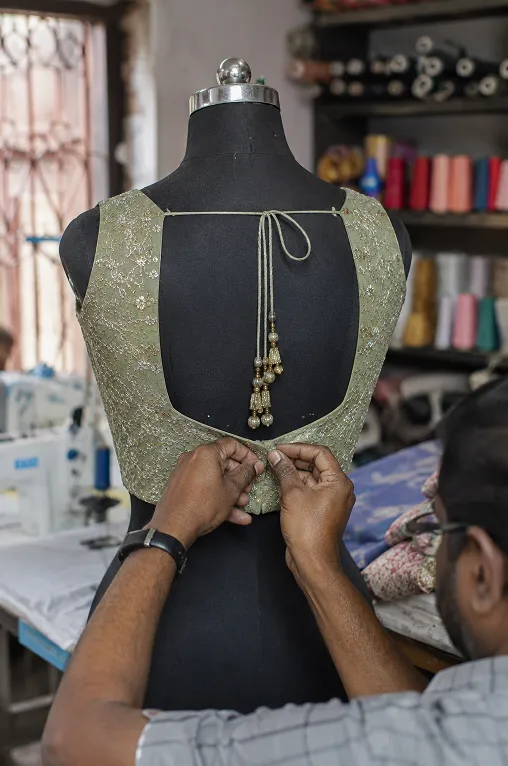 Tailor pinning the back of a saree blouse on a dress form in a textile workshop, showing precise blouse fitting and stitching details.
