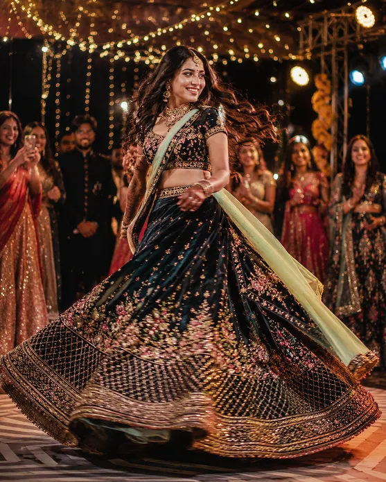 Indian woman twirling in a heavily embroidered lehenga during a sangeet celebration with festive lights and guests blurred in the background.